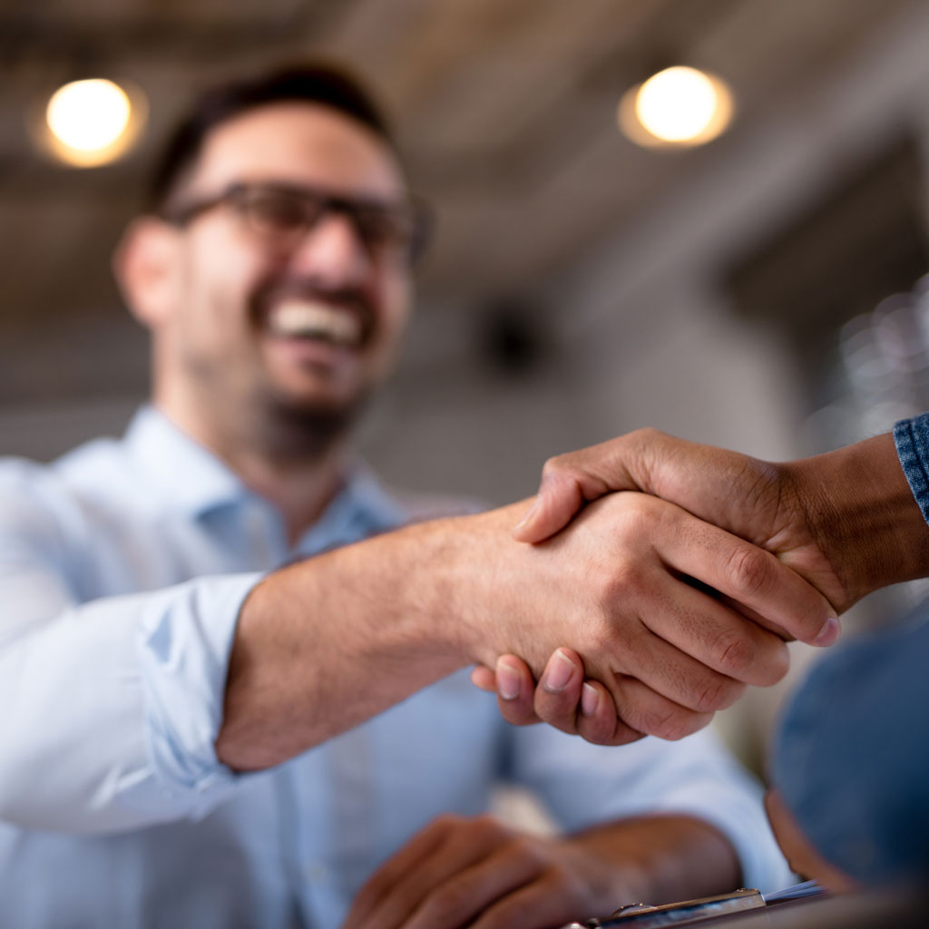 A close-up of two people shaking hands warmly. A smiling bearded man in glasses is blurred in the background, conveying a positive and friendly tone.