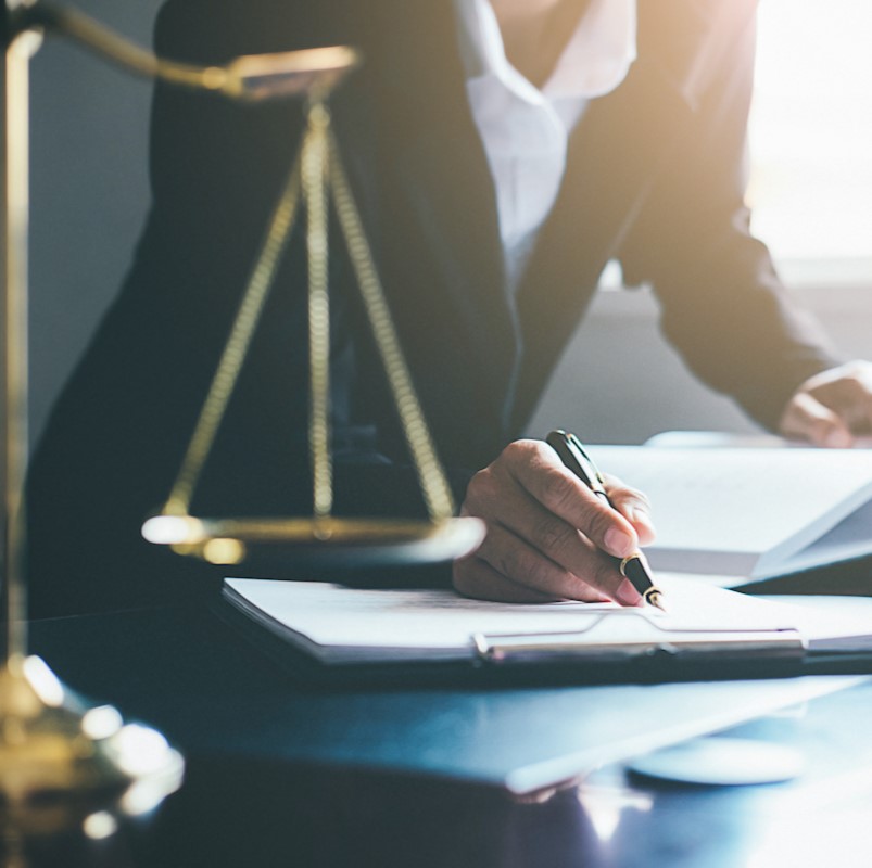 A person in a suit is writing in a notebook at a desk, with a golden balance scale in the foreground. The scene conveys a professional legal setting.