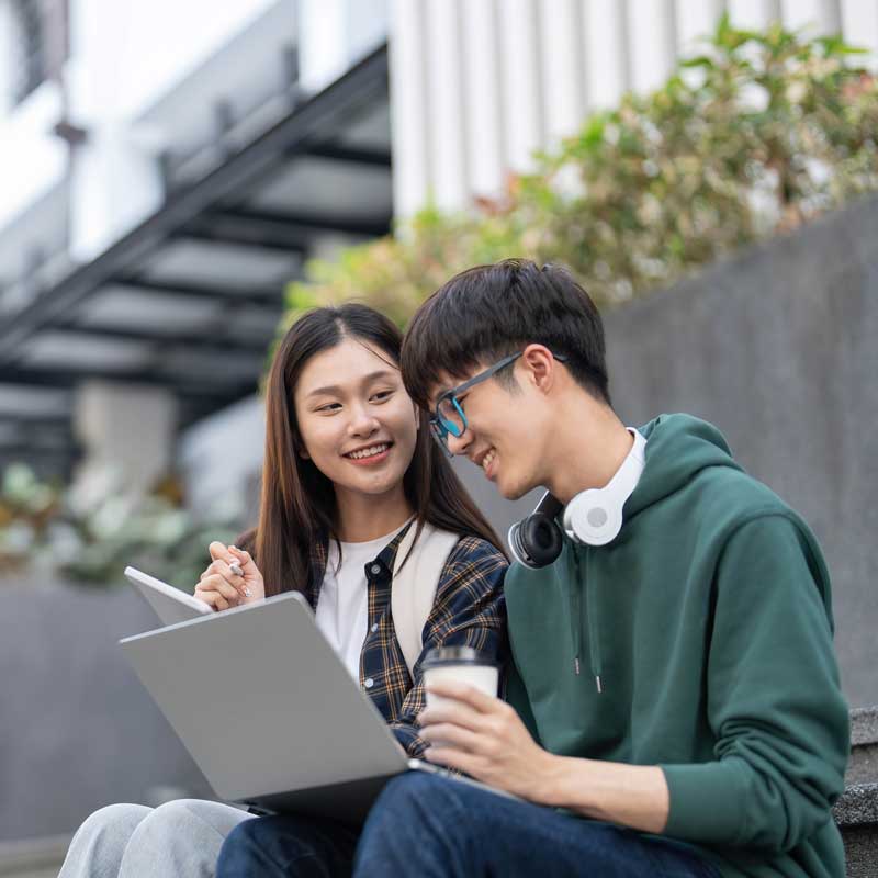 A young woman and man sit outside, smiling and looking at a laptop. The man holds a coffee cup, wearing headphones around his neck. Relaxed, cheerful atmosphere.